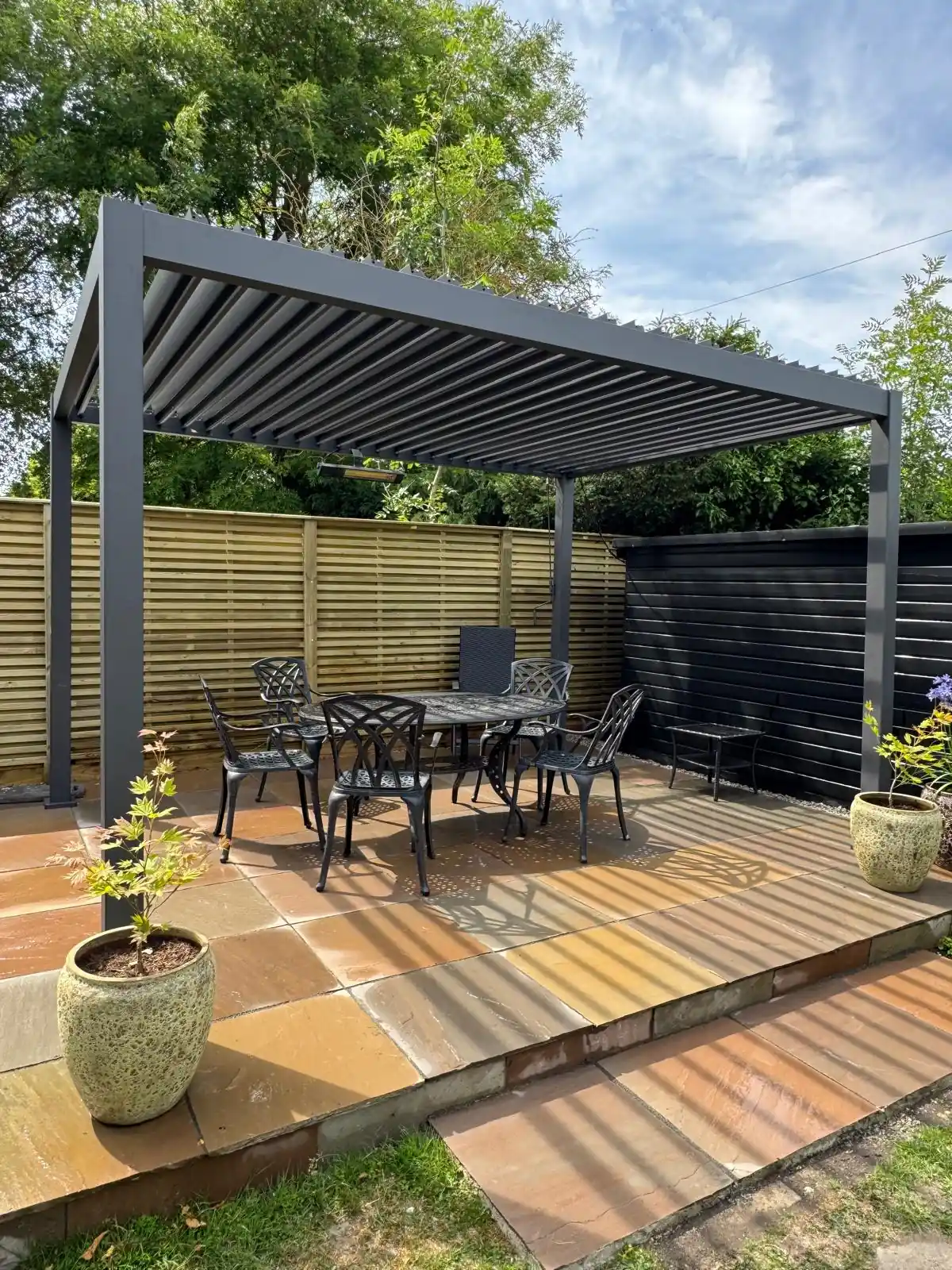 Anthracite pergola over a terracotta and sandstone patio with cast iron table and chairs, slatted and dark painted fencing behind