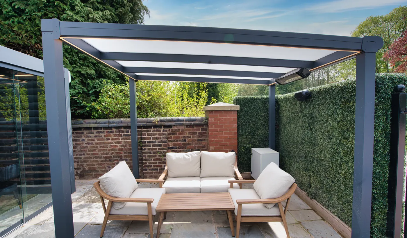 Four-post aluminium pergola sited in a garden corner between a brick wall and a clipped hedge, with lounge seating and an overhead heater