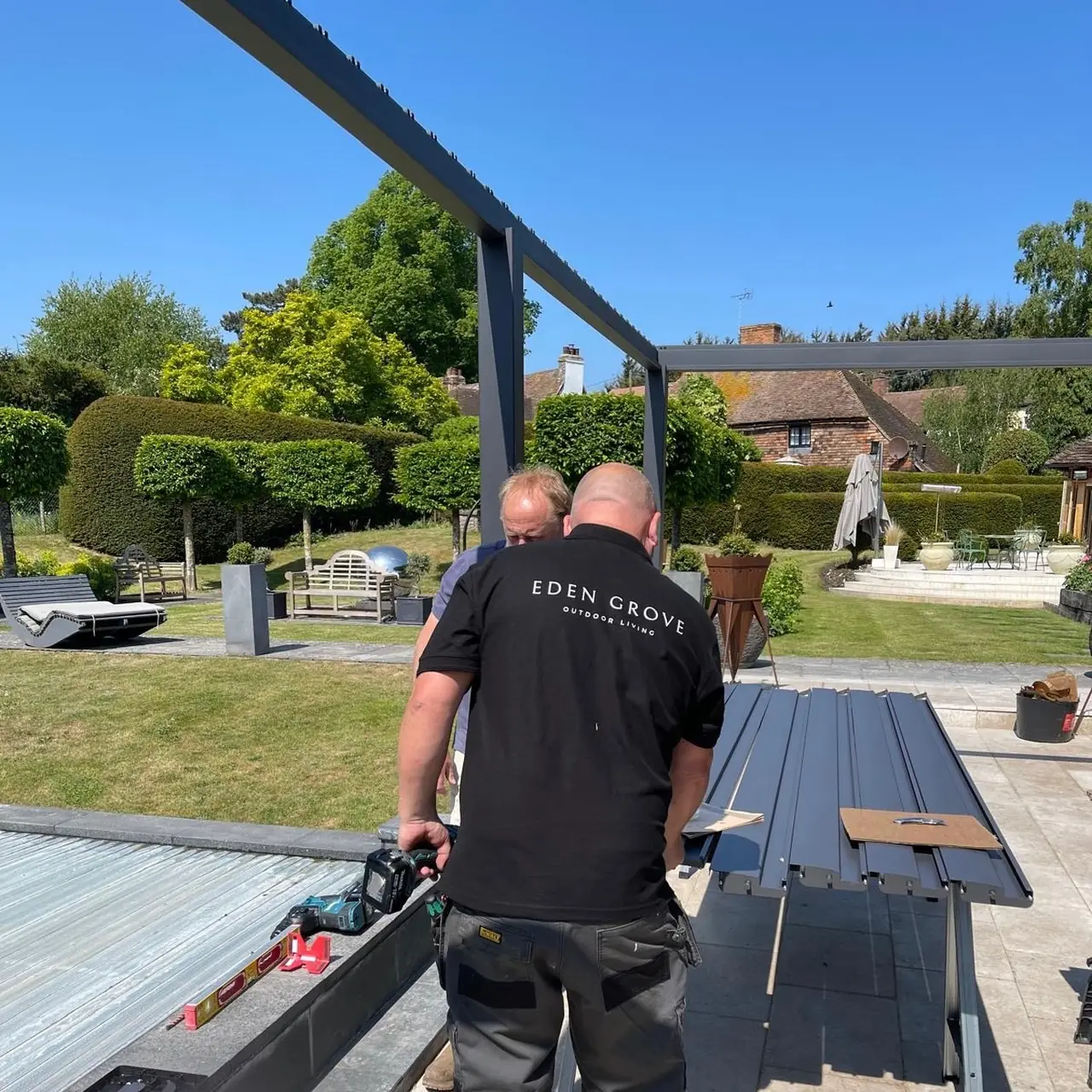 Eden Grove installer in a branded polo working on an aluminium frame, roof blades laid out on the terrace beside him