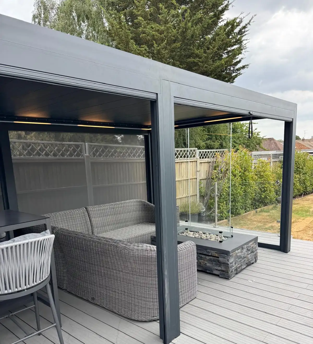 Morvelle pergola in anthracite with LED lighting integrated along the roof beams, glass side panels, a rattan corner sofa and a gas fire pit on grey composite decking