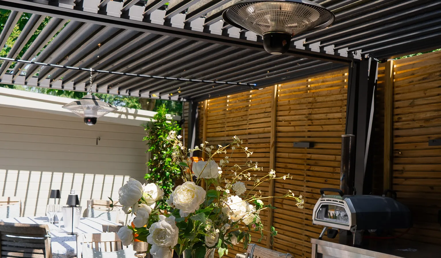 Louvred aluminium pergola over a dressed teak dining table, with a heater overhead and an outdoor kitchen against a slatted timber fence