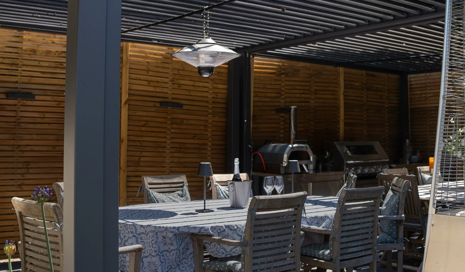 Dining table set under a louvred pergola beside a built-in outdoor kitchen and slatted timber screen