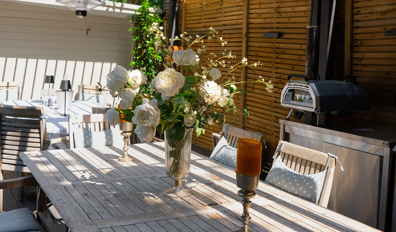 Long teak table dressed with white flowers and candles under a louvred pergola, pizza oven alongside