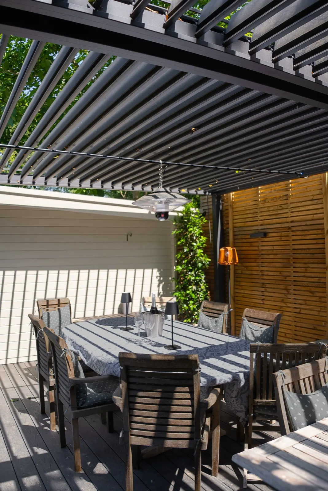 Table lamps and a hanging heater over a dining table under a louvred pergola roof