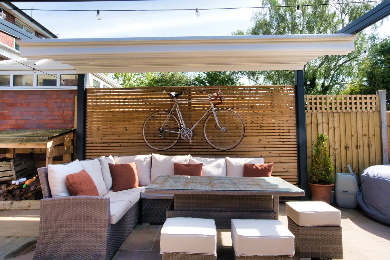Verdeca folding fabric roof retracted over a corner sofa and dining table, slatted timber screen and bicycle behind