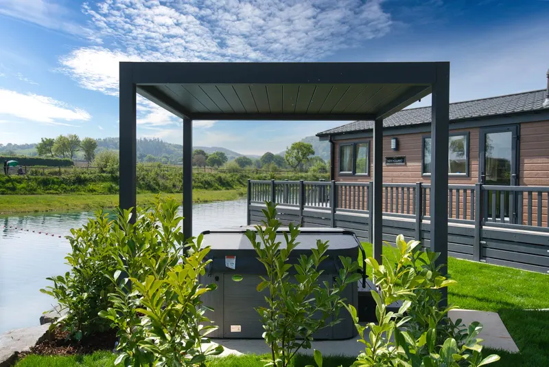 Deponti Pinela pergola over a hot tub on a lawn beside a lake and lodge