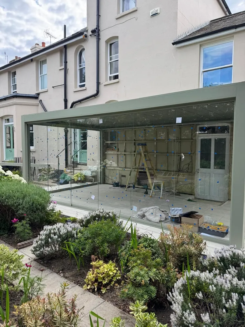 Sage green Morvelle pergola spanning the back of a cream rendered house, glass walls fitted, borders in front