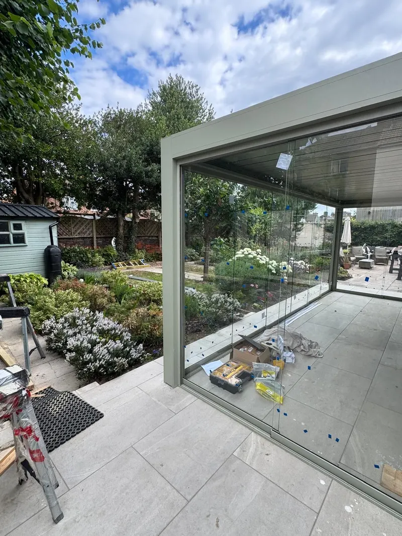 Sage green Morvelle pergola corner with frameless glass sides, porcelain paving and cottage garden beyond