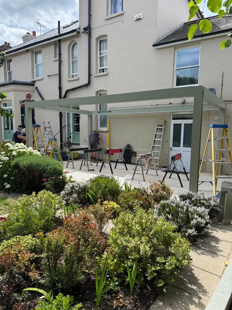 Sage green Morvelle pergola frame and roof in place against a cream house, trestles on the patio