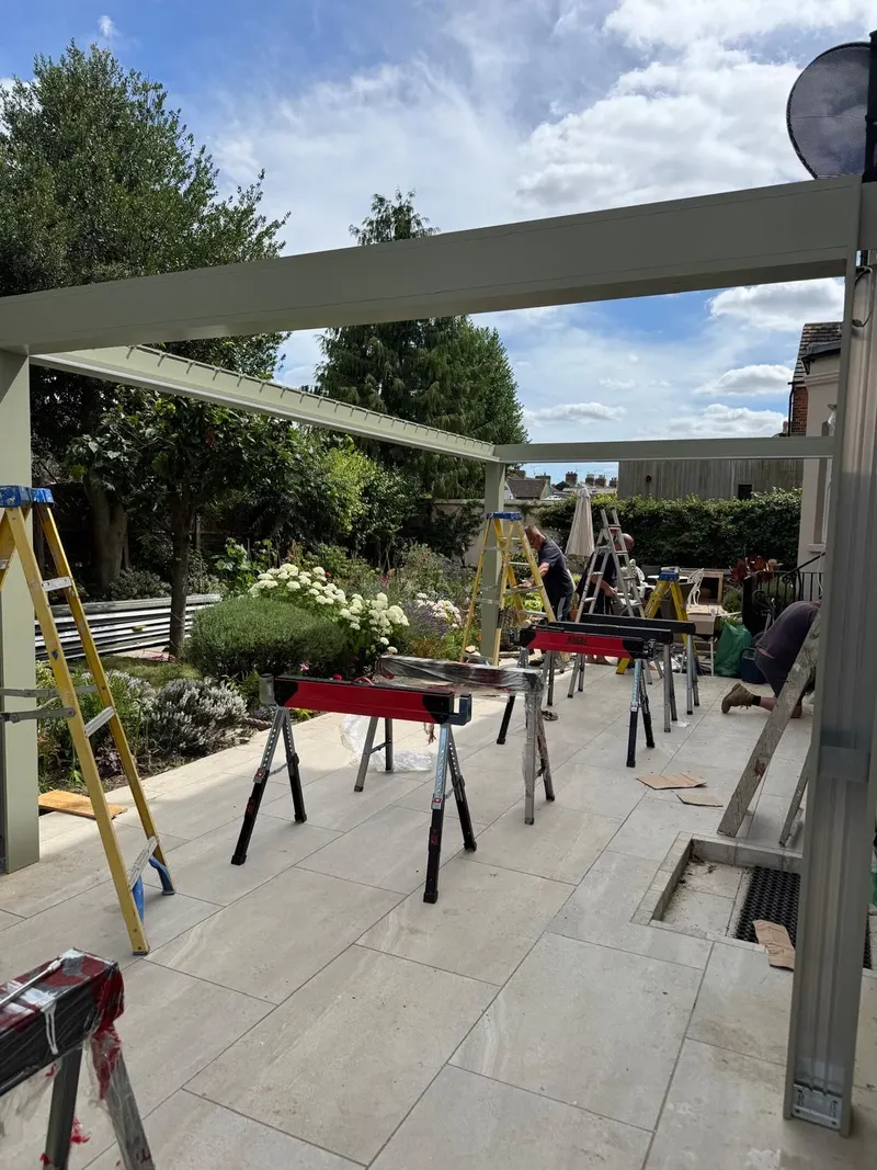 Sage green Morvelle pergola frame under a blue sky, trestles and ladders set out on the paving