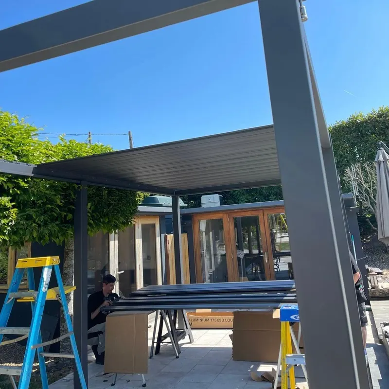Eden Titan pergola frame standing on paving mid-build, ladders and louvre blades on trestles beneath