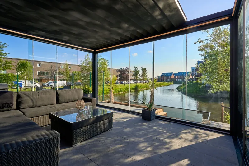 Deponti Pinela Deluxe anthracite roof enclosed with glass sliding walls, lounge seating inside and a canal beyond