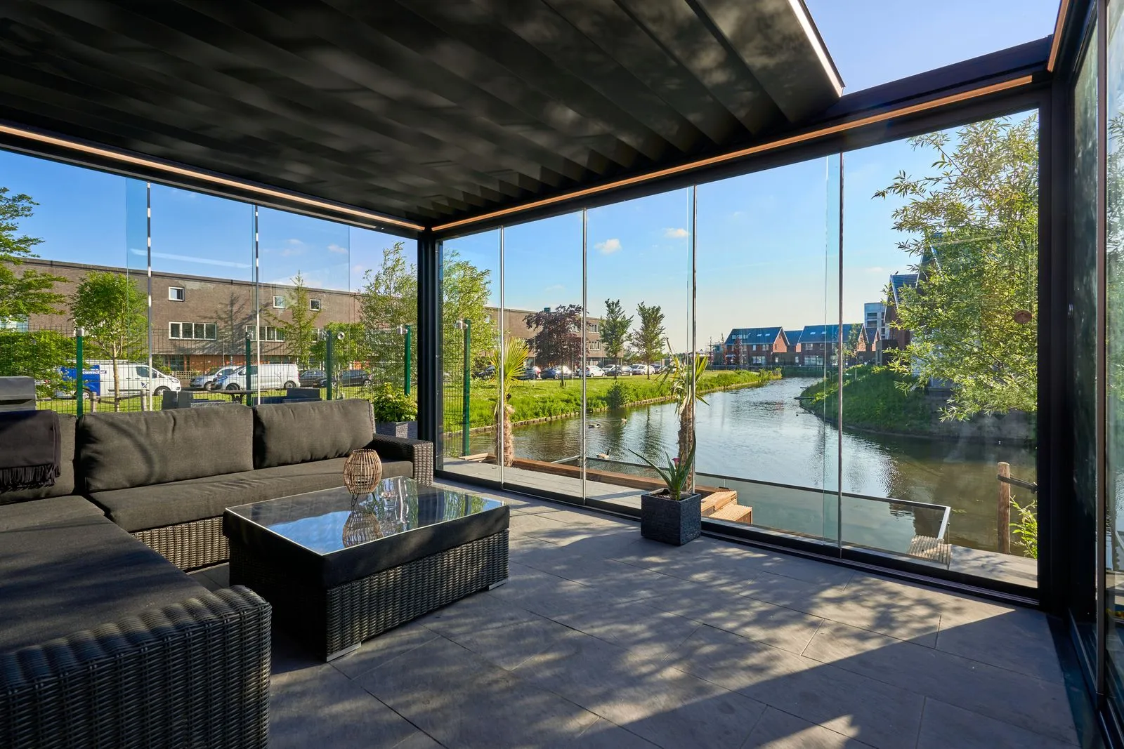 Deponti Pinela Deluxe anthracite roof enclosed with glass sliding walls, lounge seating inside and a canal beyond